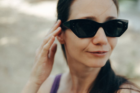 A beautiful woman in stylish sunglasses, smiling and touching her hair on a sunny day. A woman wearing fashionable black sunglasses touches her face outdoors. She has a pleasant expression.の写真素材