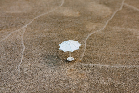 A miniature white parasol sits on textured sand, creating a simple yet captivating scene. A miniature white parasol stands on a sandy surface, possibly a beach or desert suggesting a small-scale sceneの写真素材