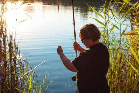 A person fishes by a lake. A woman holding fishing rod and small fish outdoors near river reeds at sunset. Concept of summer leisure, outdoor lifestyle, eco living and authentic moments in nature.の写真素材