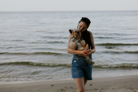 A woman and her yorkie enjoy a peaceful moment by the ocean. A woman lovingly embraces her Yorkshire terrier doggy on a sandy beach, with a serene ocean or sea backdrop, enjoying a peaceful moment.の写真素材