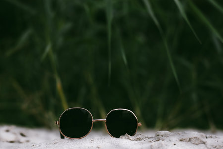 Stylish round sunglasses on sand, with a blurred green background. A pair of round sunglasses with black glasses rests on a sandy surface, with green foliage in the blurred background. Palm fond.の写真素材