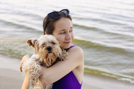 A young pretty brunette woman cuddles adorable Yorkshire terrier doggy on a sunny beach day at the...