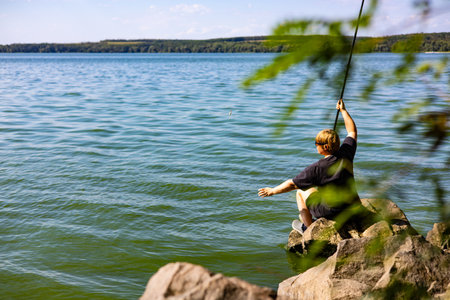 A young boy fishes by the lake on a sunny day, enjoying the outdoors and having fun. A fisherman fishing by the lake, enjoying a sunny day outdoors. Peaceful lakeside scene with fishing rod in handの写真素材
