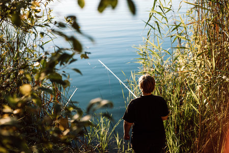 A mature woman fishing on a sunny day on a lake shore. A back view of a person fishing on a river bank, coast. The reeds and trees on a foreground and a calm water in the pond. Summer activity outdoorの写真素材