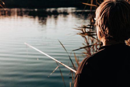 Person fishing by the lake at sunset. A serene shot of a woman viewed from the back fishing alone, silhouetted against a tranquil lake, bathed in the warm hues of the setting sun Peaceful solo fishingの写真素材