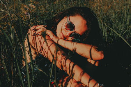 Woman relaxing in a field A woman wearing sunglasses rests amongst tall grass on a sunny day, casting shadows on her skin. Moody female portrait in summertime. Leisure and fun.の写真素材