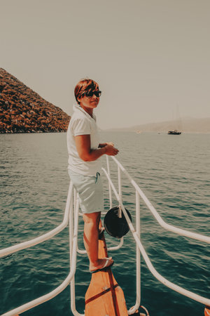 Woman on a boat enjoying a sunny day A person stands on a boat's railing, looking back at the camera with a smile, with a backdrop of sea, mountains.の写真素材
