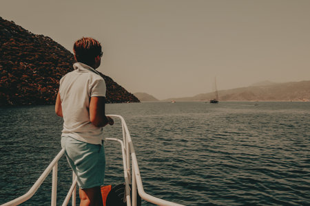 Woman on a boat enjoying a sunny day A person stands on a boat's railing, looking back at the camera with a smile, with a backdrop of sea, mountains. A person gazes out at the calm sea and islands.の写真素材