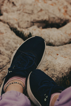 Person wearing blue sneakers and a beaded bracelet relaxing on a rocky surface. Female feet resting...