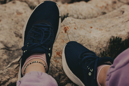 Person wearing blue sneakers and a beaded bracelet relaxing on a rocky surface. Female feet resting...