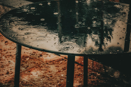 Raindrops on a round black table outdoors. Close-up shot of rain drops creating ripples on a dark table surface, with reflections of the surroundings. Cold autumnal season. Stormy weather in fall dayの写真素材