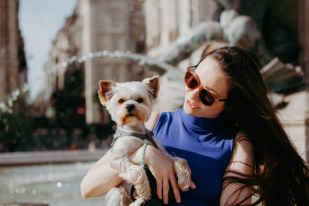 Young brunette woman holding a small dog Yorkshire terrier in front of a fountain. A girl with sunglasses holds a doggy in her arms, posing on a city street on a sunny day. A trip with a lapdog, pet.の写真素材