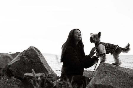 Black and white photo of a young woman with long hair gently touching her small Yorkshire terrier doggy in clothes on seaside rocks. Emotional portrait showing love connection of human and lovable petの写真素材