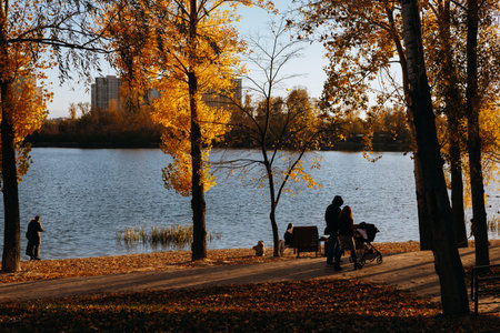 Kyiv, Ukraine 26 October 2025 Autumn park scene with a lake, trees, and people enjoying a sunny day. A beautiful autumn day in the park with a river, trees with yellow leaves Families enjoying a walk.のeditorial素材