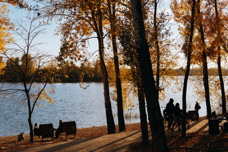 Kyiv, Ukraine 26 October 2025 Autumn park scene with a lake, trees, and people enjoying a sunny day. A beautiful autumn day in the park with a river, trees with yellow leaves Families enjoying a walk.のeditorial素材