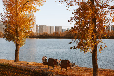 Kyiv, Ukraine 26 October 2025 Autumn scene with trees, lake, benches, and city buildings in the background. The photograph captures a serene autumn day with vibrant foliage and a tranquil waterscape.のeditorial素材