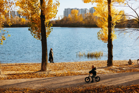 Kyiv, Ukraine 26 October 2025 Autumn scene with a child riding a bike near a lake, yellow trees. A young child enjoys a bike ride on a path near a lake with fall foliage and a person walking nearby.の写真素材
