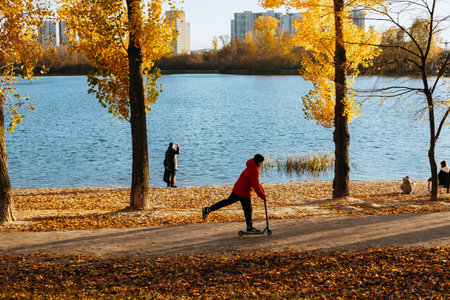 Kyiv, Ukraine 26 October 2025 Autumn scene with a child riding a scooter near a lake with gold trees. A kid rides a scooter along a path near a river surrounded by trees with yellow leaves Fall seasonのeditorial素材