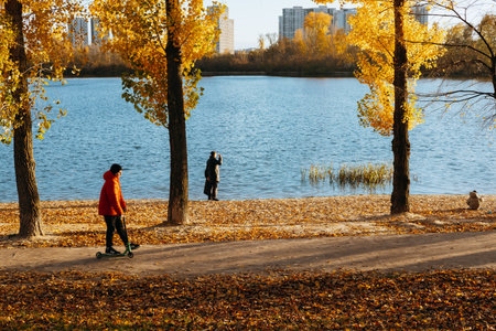 Kyiv, Ukraine 26 October 2025 Autumn scene with a child riding a scooter near a lake with gold trees. A kid rides a scooter along a path near a river surrounded by trees with yellow leaves Fall seasonのeditorial素材