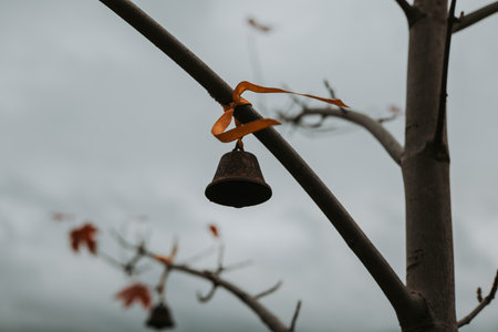 Rustic bell tied to a tree branch with a ribbon against a cloudy sky. A small, weathered bell hangs from a tree branch. A symbol of protection from evil spirits, a good luck charm. Moody autumnal day.の写真素材