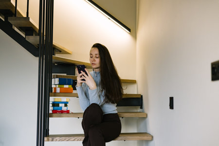 Young brunette woman using smartphone while sitting on modern wooden stairs at home, cozy minimalist...