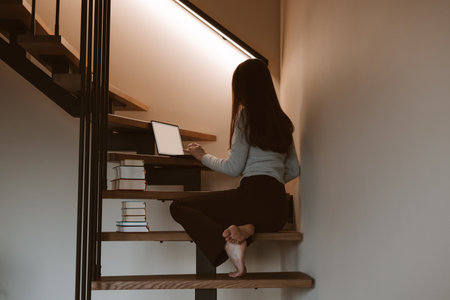 Young woman working on laptop at home office sitting on wooden stairs, cozy minimalist interior, remote work and creative freelance lifestyle concept, calm productivity and modern slow living vibe.の写真素材