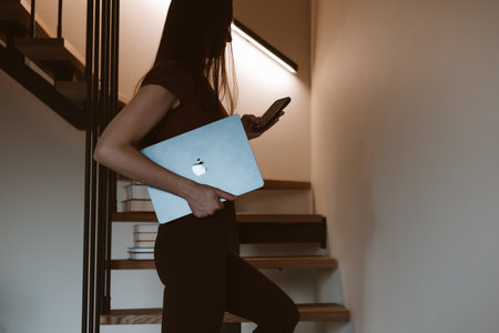 KYIV, Ukraine 1 November 2025 Woman using phone while walking up stairs with Apple MacBook laptop. A businesswoman is holding a laptop and smartphoneのeditorial素材