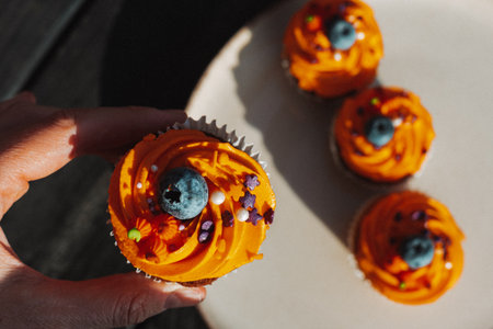 Delicious Halloween cupcake with orange frosting and blueberry held by a hand. A hand holds a cupcake with orange frosting, a blueberry, and decorative sprinkles, with other cupcakes in background.の写真素材
