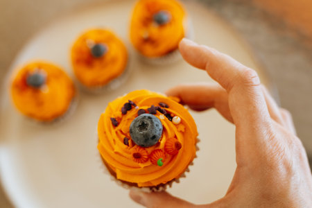 Delicious Halloween cupcake with orange frosting and blueberry held by a hand. A hand holds a cupcake with orange frosting, a blueberry, and decorative sprinkles, with other cupcakes in background.の写真素材