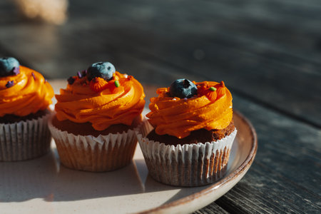 Delicious cupcakes with orange frosting and blueberry toppings on a plate on a rustic dark wooden table. These tasty sweets and treats are perfect for a special occasion like Halloween holiday.の写真素材