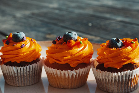 Delicious cupcakes with orange frosting and blueberry toppings on a plate on a rustic dark wooden table. These tasty sweets and treats are perfect for a special occasion like Halloween holiday.の写真素材
