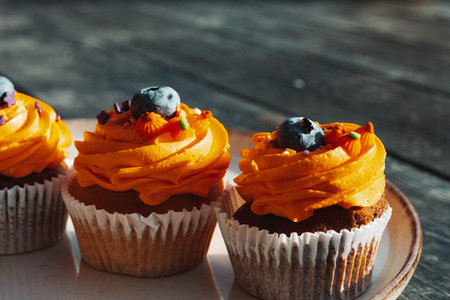 Delicious cupcakes with orange frosting and blueberry toppings on a plate on a rustic dark wooden table. These tasty sweets and treats are perfect for a special occasion like Halloween holiday.の写真素材