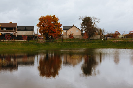 KYIV, Ukraine 1 November 2025 Autumn suburban landscape with houses, colourful trees, reflective lake and cloudy sky. The tranquil scene captures the beauty of fall foliage and the serene reflection.のeditorial素材