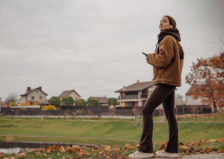 Stylish young woman walking outdoors in autumn park with smartphone, cozy brown outfit, calm mindful lifestyle, slow living and digital minimalism concept. Digital balance and slow living outdoor.の写真素材