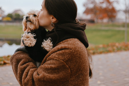 Woman in a warm brown coat cuddling and kissing her small Yorkshire Terrier outdoors in autumn park. Cozy fall vibes, love for pets and friendship between human and dog A girl kissing her Yorkie doggyの写真素材