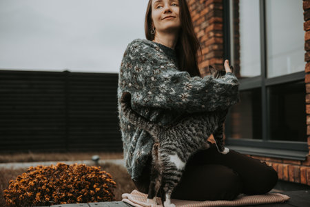 Warm autumn moment with a young woman sitting outside brick house, playing with a cat while enjoying a calm cozy day. Relaxed lifestyle scene showing love for pets, slow living. Comfort, home warmth.の写真素材