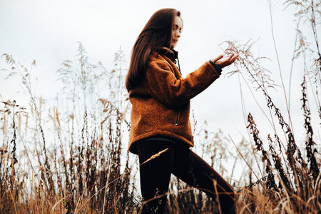 A girl in a warm jacket touching tall grass while walking in a field on a cloudy day. A young...