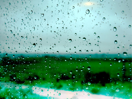 Raindrops on glass with a blurred green field and glowy gray sky in the background. The image captures water droplets on a window, with a soft focus on the green landscape beyond. Rainy season.の写真素材