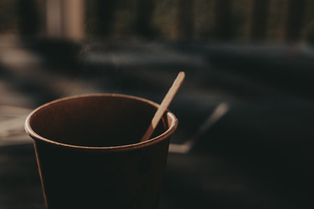 Steaming coffee in a paper cup with a wooden stirrer, close-up shot. A warm cup of takeaway coffee sits on a table, with steam rising from the top, creating a cozy atmosphere. Morning lifestyle.の写真素材
