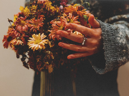 A woman's hand with a massive silver ring on a finger in a gray knitted sweater, holding a lush autumn bouquet of chrysanthemums in warm tones. The orange, yellow flowers create a cozy, autumnal sceneの写真素材