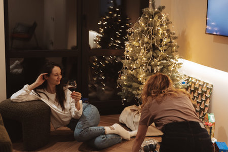 Authentic holiday moment of friends decorating a Christmas tree for 2026. Warm lights, cozy home interior, gifts, relaxed atmosphere, real-life celebration vibes capturing modern festive togetherness.の写真素材