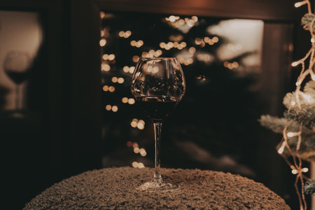 Elegant wine glass with red wine, festive lights in the background. Soft lights creating a warm and inviting atmosphere. Celebration New Year 2026 at home. Dark moody scene. Christmas garland lights.の写真素材