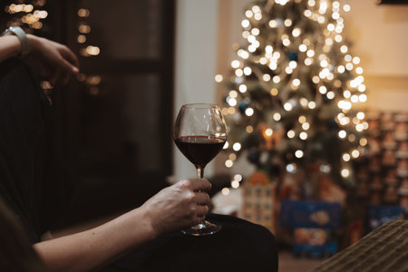 Cozy Christmas evening scene, a person holds a glass of rich red wine against a background of a decorated festive tree with sparkling bokeh lights. Ideal for holiday, winter, and celebration concepts.の写真素材