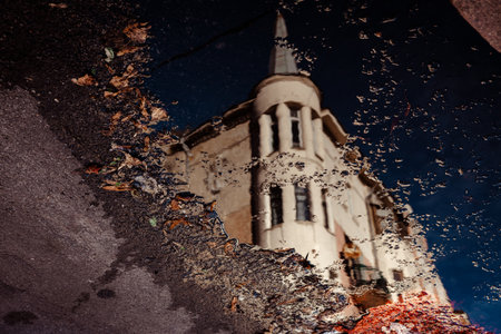 A striking reflection of a unique, old urban building in a puddle after rain Moody high-contrast image captures architectural details and the sky in the water's surface, highlighted by a splash of redの写真素材