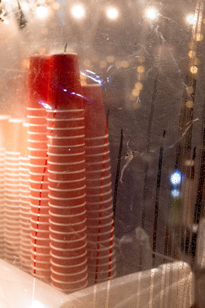 Stacked red and white paper coffee cups behind a wet plastic film. Warm, blurred bokeh lights in the...