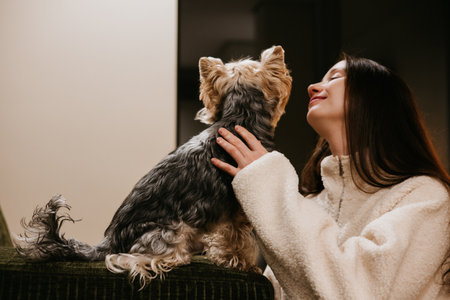 Cozy young woman in a fluffy white sweater looking at and petting a small Yorkshire Terrier dog. This authentic lifestyle photo captures a tender moment of care and love between a pet owner and doggyの写真素材