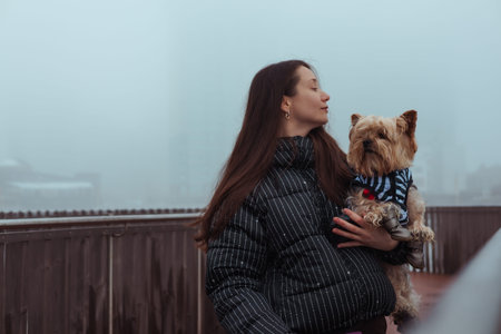 A beautiful young woman in a stylish puffer jacket holding a cute Yorkshire Terrier dog outdoors on a misty winter urban day. This lifestyle portrait captures the warm bond between owner and pet.の写真素材