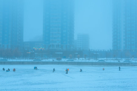 KYIV, UKRAINE - FEBRUARY 13, 2026: Ice fishermen sit on the frozen river surface in the city during a foggy day. Urban winter landscape with buildingsのeditorial素材