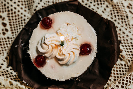 Top view of a small white birthday cake with whipped cream, cherries, and a burning candle on a lace...