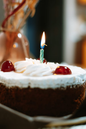 Chocolate birthday cake with whipped cream, cherries, and a burning candle on a table. Moody vintage...
